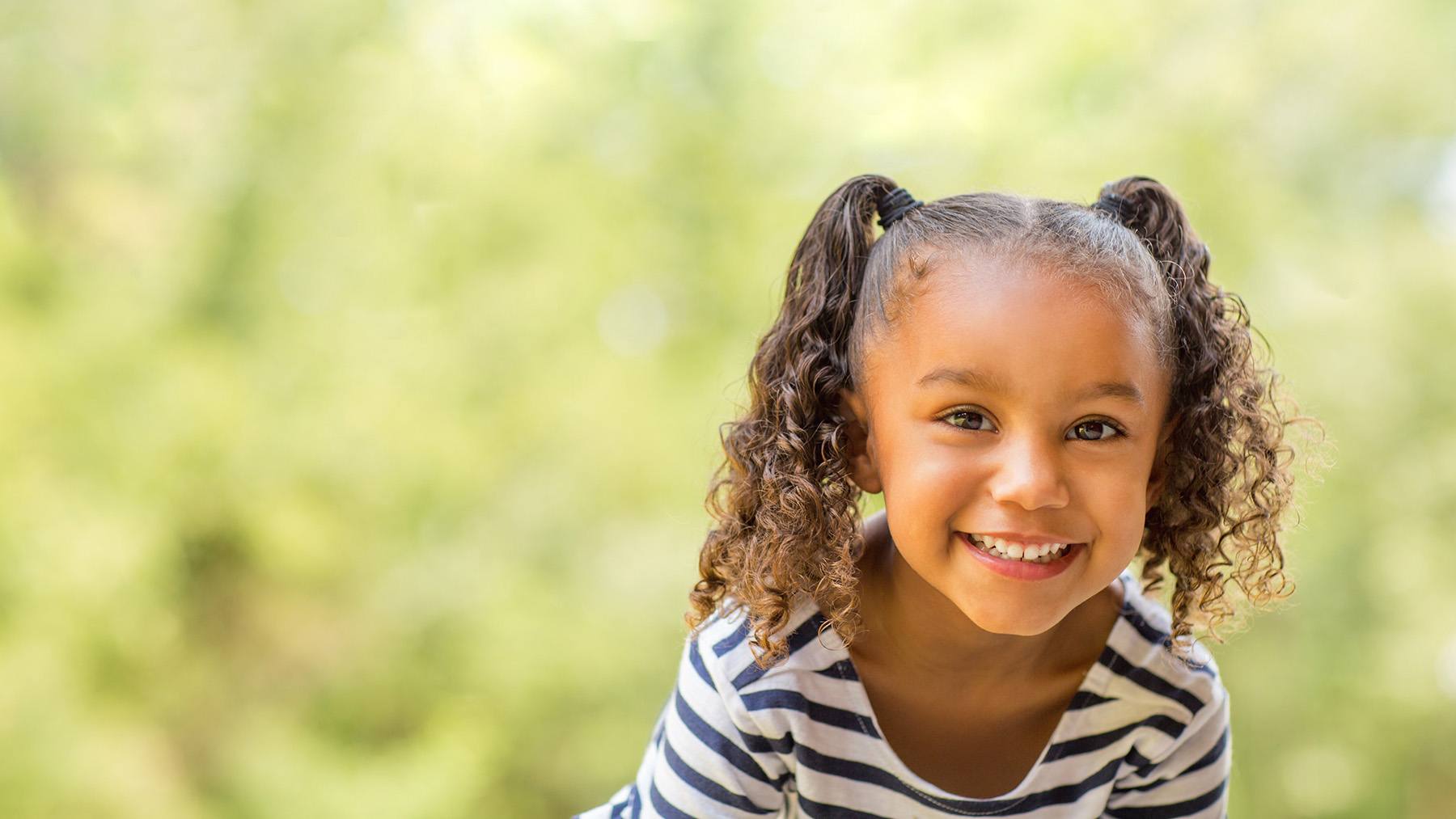 A young girl with curly hair and a joyful expression, smiling at the camera while sitting outdoors.
