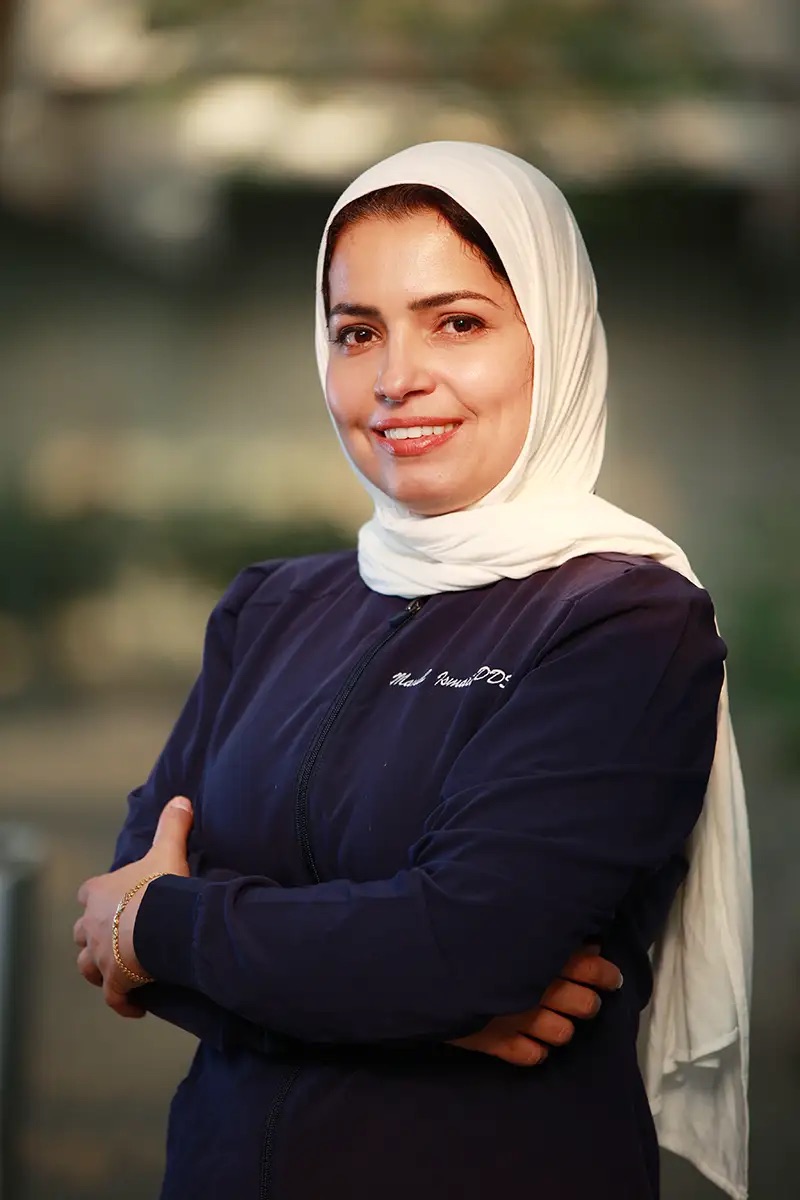 The image features a smiling woman wearing a hijab, standing with her arms crossed against a blurred background, likely posing for a formal portrait.